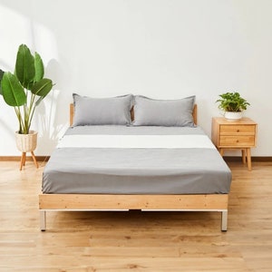 May include: A wooden bed frame with a gray mattress and two gray pillows. The bed is in a room with a white wall and wooden floors. There is a small plant on a wooden nightstand to the right of the bed.