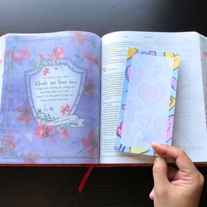 May include: An open Bible with a blue and pink heart-shaped bookmark. The bookmark has a white background with a cross and hearts drawn on it. The Bible has a floral design on the cover.