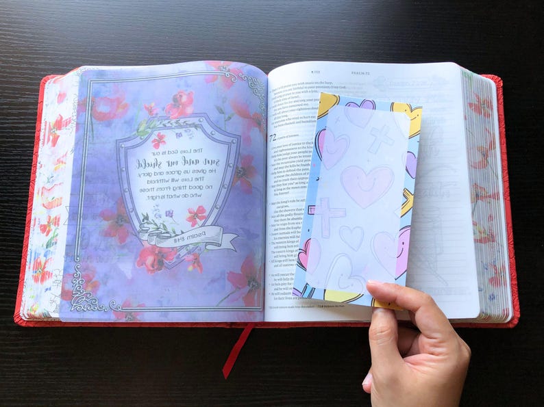 May include: An open Bible with a blue and pink heart-shaped bookmark. The bookmark has a white background with a cross and hearts drawn on it. The Bible has a floral design on the cover.