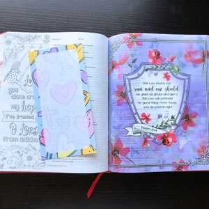 May include: An open Bible with a blue and white bookmark with hearts. The page is open to Psalm 84, which reads "The Lord God is our sun and our shield. He gives us grace and glory. The Lord will withhold no good thing from those who do what is right."