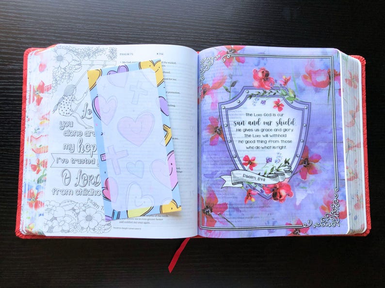 May include: An open Bible with a blue and white bookmark with hearts. The page is open to Psalm 84, which reads "The Lord God is our sun and our shield. He gives us grace and glory. The Lord will withhold no good thing from those who do what is right."