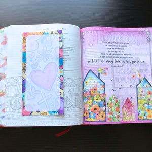 May include: An open Bible with a pink and white floral design on the right page. The left page has a colorful bookmark with a heart and cross design. The text on the right page reads "He has torn us to pieces, now He will bandage our wounds, so that we may live in His presence.  Hosea 6:1-2.  Look and be willing".