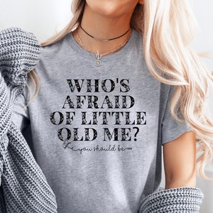 May include: A gray t-shirt with the text "Who's Afraid of Little Old Me?" in black with a decorative floral design. The text "You should be..." is written in cursive below the main text.