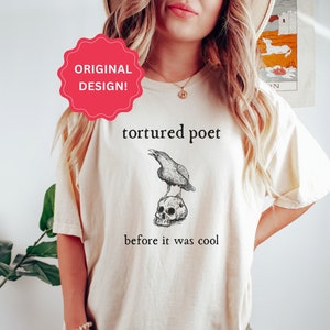 May include: Cream-colored t-shirt featuring a black graphic of a raven perched atop a skull, with the words "tortured poet" and "before it was cool" printed in black. The shirt has a relaxed fit.