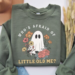 May include: An olive green sweatshirt featuring a ghost and floral graphic. The text reads "WHO'S AFRAID OF LITTLE OLD ME?" The design has a retro style. The sweatshirt is a crew neck style.