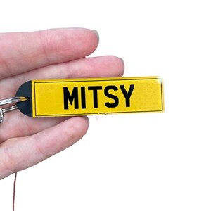 May include: A yellow keyring with a black border and the word "MITSY" printed in black letters.