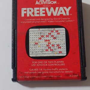 May include: A red and black Atari game cartridge for the game Freeway. The cartridge has a black label with white text that reads "ACTIVISION PRESENTS FREEWAY". The label also includes the text "Conceived and designed by David Crane for enjoyment on your Atari Video Computer System."