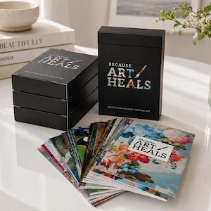 Op de afbeelding: Zwarte kaartenboxen met de tekst "BECAUSE ART HEALS" en "REFLECTIONS ON GRIEF THROUGH ART". De kaarten hebben kleurrijke bloemenkunst. Een stapel van drie dozen is zichtbaar.
