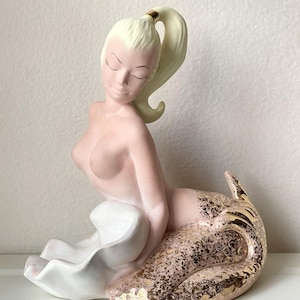May include: A ceramic mermaid figurine with pale pink skin, a blonde ponytail tied with a gold band, and a closed-eyed expression. The mermaid rests on a white shell-like base, and her tail features a textured pink finish with gold leaf accents.
