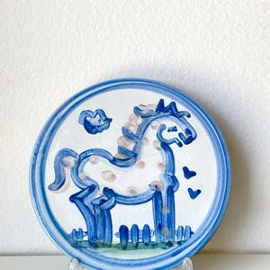 Puede incluir: Un plato redondo de cerámica con un caballo azul pintado a mano con manchas de color marrón claro. El diseño incluye una nube azul, dos pequeños corazones azules y una base de hierba verde. El plato tiene un borde azul grueso y un soporte.