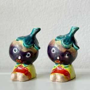 Puede incluir: Dos saleros y pimenteros de cerámica con forma de personajes de dibujos animados. Los saleros están pintados para parecer berenjenas con hojas verdes en la parte superior. Los personajes tienen caras negras con ojos blancos y bocas rojas. Los saleros están sobre una superficie blanca.