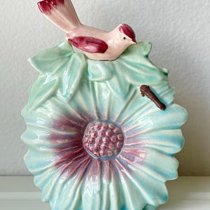 May include: A ceramic decorative object shaped like a large, light blue flower with a textured, dark pink center. A small, light pink bird with dark red wing and tail accents is perched on top of the petals. A small brown twig is attached to the side.
