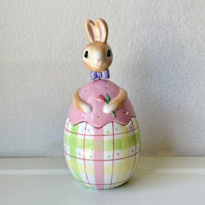 Peut inclure: Une figurine de lapin de Pâques en céramique tenant une fleur rose. Le lapin porte un nœud papillon violet et est assis dans un œuf à carreaux rose et vert.