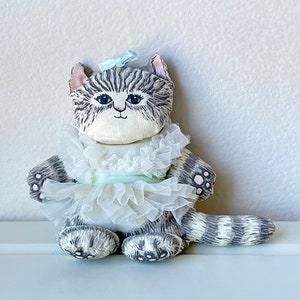 Puede incluir: Un gato de peluche gris y blanco con una falda de tul blanca y un lazo de cinta azul claro. El gato tiene almohadillas de patas rosas y una cara bordada en negro.