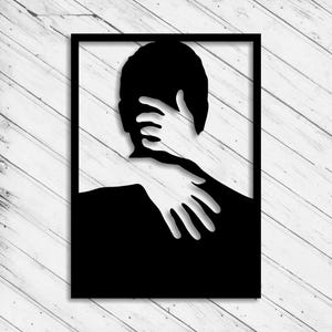 May include: Black metal wall art depicting a silhouette of a person being embraced. The hands of the person embracing are in white, contrasting with the black figure. The art is framed in a black rectangle.