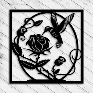 May include: Black metal wall art featuring a hummingbird hovering near a rose. The design includes a circular frame with floral elements and a square border. The hummingbird is depicted with detailed wing and feather patterns.