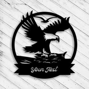 May include: Black metal wall art featuring an eagle in flight, set within a circular frame. The eagle is depicted with outstretched wings, and the text "Your Text" is displayed on a banner below. Two birds are flying above the eagle.