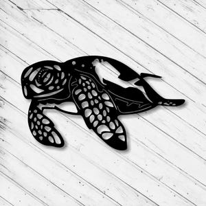 Op de afbeelding: Zwarte metalen wandkunst van een zeeschildpad. De schildpad is in profiel afgebeeld met gedetailleerde uitsparingen op de schild en vinnen. De achtergrond is een wit houten oppervlak.