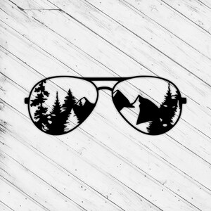 May include: Black aviator sunglasses with a mountain and forest scene reflected in the lenses. The sunglasses are set against a white wooden background. The lenses show a silhouette of trees and mountains.