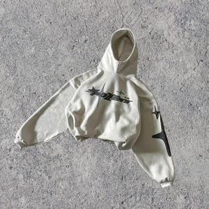 Puede incluir: Una sudadera con capucha gris claro con un gr&aacute;fico negro de una estrella y el texto "XOLVER" en la parte delantera. La sudadera tiene gr&aacute;ficos de estrellas negras en las mangas.