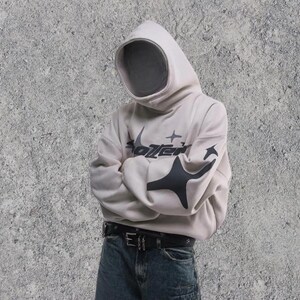 Puede incluir: Una sudadera con capucha blanca con un dise&ntilde;o gr&aacute;fico negro que dice "Sotzen" con estrellas y una forma geom&eacute;trica negra. La sudadera se lleva sobre un vaquero azul con un cintur&oacute;n negro.
