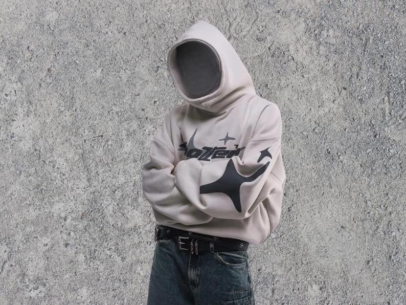 Puede incluir: Una sudadera con capucha blanca con un dise&ntilde;o gr&aacute;fico negro que dice "Sotzen" con estrellas y una forma geom&eacute;trica negra. La sudadera se lleva sobre un vaquero azul con un cintur&oacute;n negro.