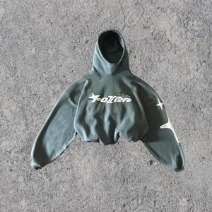 Puede incluir: Una sudadera con capucha verde recortada con una estrella blanca y el texto "Fallen" en la parte delantera. La sudadera tiene dise&ntilde;os de estrellas blancas en las mangas.