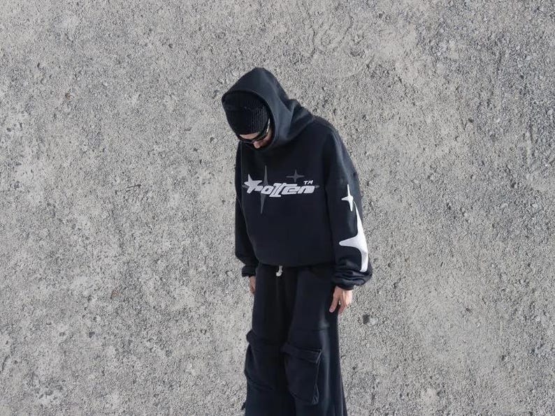Puede incluir: Una sudadera con capucha negra con una estrella blanca y la palabra "Rotten" impresa en la parte delantera. La sudadera tiene dise&ntilde;os de estrellas blancas en las mangas.