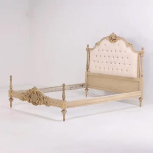 May include: A beige and gold ornate bed frame with a tufted headboard. The headboard is upholstered in a cream-colored fabric and features a decorative gold crest. The bed frame has detailed carvings and a classic design.
