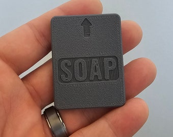 Può includere: Un piccolo dispenser di sapone rettangolare grigio con la parola "SOAP" stampata su di esso. Una freccia che punta verso l'alto è stampata sopra la parola.