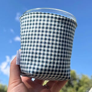 May include: A glass cup sleeve featuring a small-scale navy blue and white gingham check pattern. The fabric sleeve is cylindrical and includes a small white rectangular fabric tag on the side with black text printed on the surface.