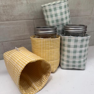 May include: Three quilted fabric sleeves for mason jars, featuring a gingham pattern in yellow and sage green. Two sleeves contain glass jars with silver metal lids, while one yellow sleeve sits empty in the foreground, showing its interior.