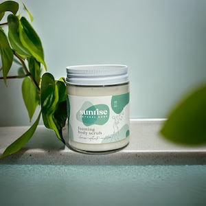 May include: A white jar with a green and white label that says "sunrise natural care" and "foaming body scrub". The jar is sitting on a light gray surface.