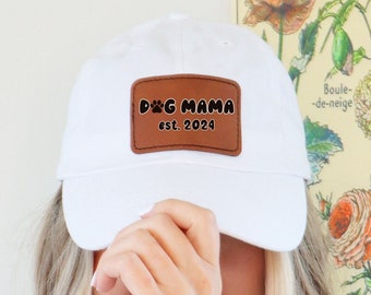 May include: A white baseball cap with a brown leather patch. The patch features the text "DOG MAMA est. 2024" in black. The cap is a classic style, suitable for casual wear. The image is set against a floral background.