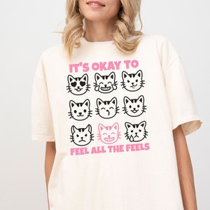Puede incluir: Una camiseta blanca de gran tamaño con un gráfico de nueve caras de gato con diferentes expresiones. El texto "It's okay to feel all the feels" está impreso debajo de las caras de gato. La camiseta se muestra en una modelo que lleva pantalones cortos negros. El color de la camiseta es marfil.
