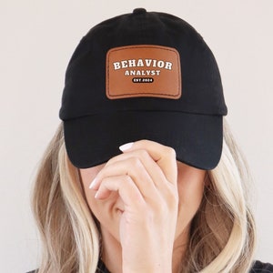 以下が含まれることがあります： 「Behavior Analyst Est. 2023」と書かれた茶色の革のパッチが付いた黒い野球帽。