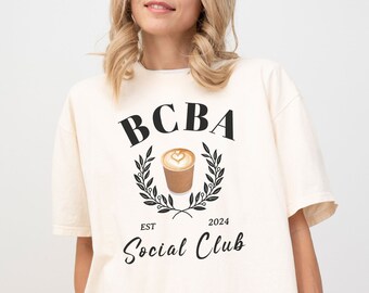 May include: A white t-shirt with a black graphic design that says "BCBA Social Club" with a coffee cup and a laurel wreath. The text "EST 2024" is below the graphic. The t-shirt is on a model wearing black shorts.