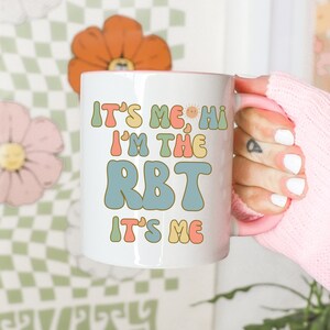 Könnte beinhalten: Weiße Keramiktasse mit dem Text "IT'S ME, HI I'M THE RBT IT'S ME" in Retro-Pastellfarben. Die Tasse wird von einer Person gehalten, die einen rosa Pullover trägt. Dekorative Blumenaufkleber und ein gerahmter Blumendruck befinden sich im Hintergrund.