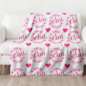 以下が含まれることがあります： ピンクのハートと円が繰り返し描かれた白いフリースの毛布。円には「Come On Beba Let's Go Anywhere」という文字が書かれています。