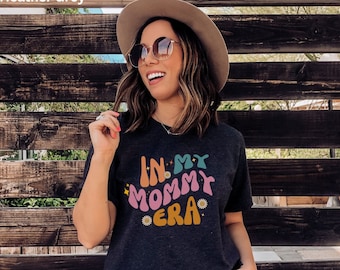 May include: A dark heather grey t-shirt with the text "In My Mommy Era" in colorful, retro-style lettering. The shirt features a relaxed fit and short sleeves.