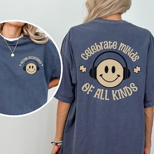 Puede incluir: Una camiseta de gran tamaño de color azul oscuro con el texto "Celebrate minds of all kinds" impreso en la espalda. El texto es de color marrón claro y presenta una cara sonriente con auriculares y piezas de rompecabezas.