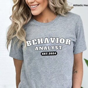 Może przedstawiać: Szary t-shirt z nadrukiem "BEHAVIOR ANALYST EST. 2024" w kolorze białym.