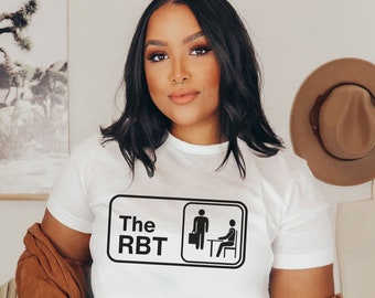 May include: White t-shirt with a black graphic of a person standing and a person sitting at a desk with the text "The RBT" above them.