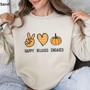 Puede incluir: Una sudadera beige con un signo de paz, un corazón y una calabaza. El texto "Happy Relaxed Engaged" está impreso debajo del gráfico.