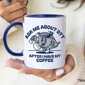 Puede incluir: Taza de cerámica blanca con borde azul. La taza presenta una ilustración de dibujos animados de una taza de café con cara, sosteniendo un lápiz y un rollo. El texto en la taza dice "Ask me about DTT after I have my coffee".