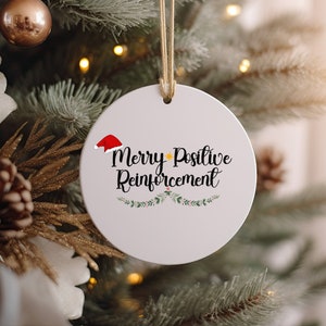 May include: White round Christmas ornament with a red Santa hat, a yellow star, and the text "Merry Positive Reinforcement" in black cursive lettering. Green and white floral accents surround the text.