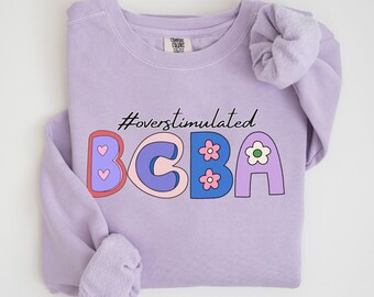 May include: A lavender sweatshirt with the text "#overstimulated BCBA" in colorful, cartoon-style letters. The letters are decorated with hearts and flowers.