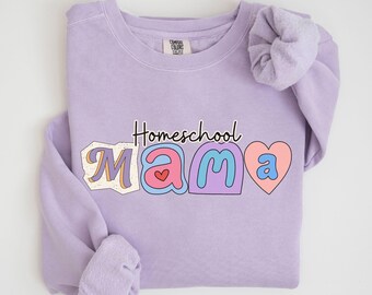 Puede incluir: Una sudadera de color lavanda con el texto "Homeschool Mama" en letras de colores con un diseño de corazón. La sudadera tiene cuello redondo y mangas largas.