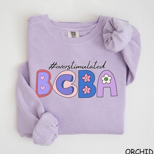 Peut inclure: Un sweat-shirt lavande avec le texte "#overstimulated BCBA" en lettres colorées de style dessin animé. Les lettres sont décorées de cœurs et de fleurs.
