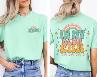May include: A light green t-shirt with a rainbow and the text "In My BCBA Era" on the back. The front of the shirt has the name "Kendra" in a retro font.